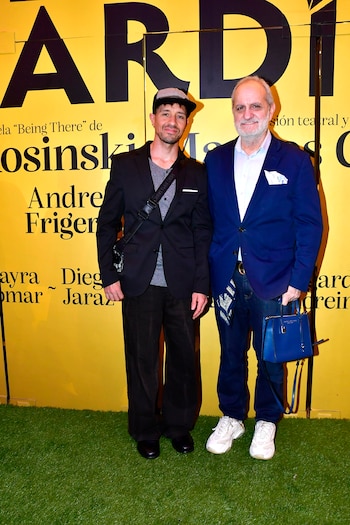 Dos hombres de pie sonríen ante un fondo amarillo con texto negro y césped artificial. Uno lleva gorra, traje negro y bandolera; el otro, saco azul y jeans