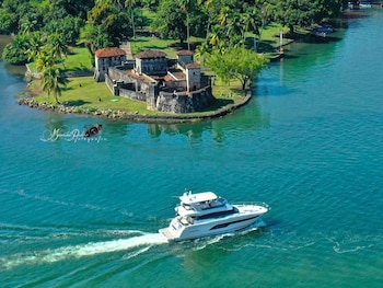 Vista aérea de un yate blanco navegando en aguas turquesas del Río Dulce, junto a la Fortaleza de San Felipe de Lara y densa vegetación verde en la orilla