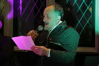 Hombre con barba canosa, vestido con chaqueta oscura, habla en un micrófono plateado, sujetando un papel rosa bajo luces moradas y verdes