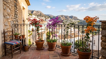 Balcón de piedra con barandilla de hierro, cuatro macetas con buganvillas de colores, una silla de madera y vista a un pueblo y montañas.