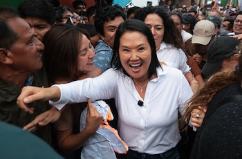 Keiko Fujimori, vestida con camisa blanca, sonríe y extiende un brazo hacia una multitud de personas que la rodean, algunas sonrientes