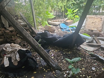 Vista de un área exterior descuidada con escombros, tablas de madera rotas, ladrillos apilados, vegetación densa y una manta negra en el suelo