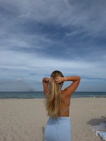 Una mujer rubia de espaldas en la playa, con las manos en el cabello. Viste una prenda clara de dos piezas y observa el mar bajo un cielo nublado