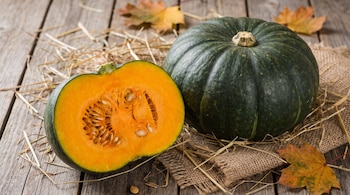 Una calabaza verde entera, una mitad de calabaza verde con pulpa naranja y semillas, heno, arpillera y hojas secas sobre una superficie de madera.