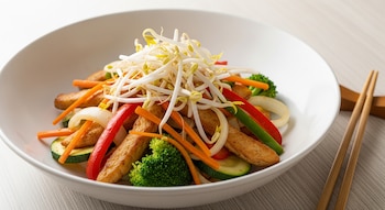 Un plato hondo blanco lleno de chop suey de pollo con tiras doradas, zanahoria, morrón rojo, cebolla, zucchini, brócoli y brotes de soja, sobre una mesa de madera clara con palitos chinos al lado.