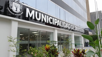 Fachada de un edificio moderno con ventanas de vidrio y la inscripción "Municipalidad de San Isidro", con el escudo local. Plantas en primer plano