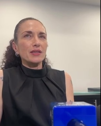 Valeria Sirgo con blusa negra sin mangas y cabello recogido habla frente a un micrófono azul