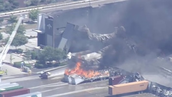 Vista aérea de un gran incendio: un tráiler de camión arde intensamente con denso humo negro elevándose junto a un edificio y contenedores de carga