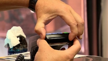 Primer plano de dos manos sosteniendo un dispositivo electrónico negro para clonar tarjetas, con detalles en azul y verde. Al fondo, un objeto borroso