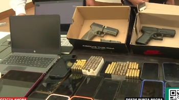 Una mesa oscura con dos pistolas en cajas de cartón, múltiples cargadores de municiones, varios teléfonos inteligentes y una laptop abierta con pantalla apagada