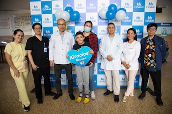 Un niño sostiene un cartel azul con forma de corazón junto a varios médicos frente a un panel con globos