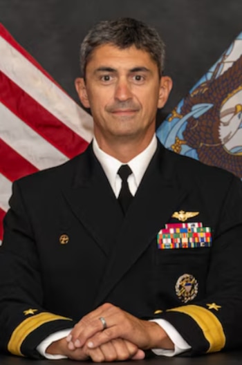 Retrato de un hombre de cabello oscuro y canoso con uniforme naval oscuro, insignias, condecoraciones y galones dorados. Fondo con bandera de Estados Unidos