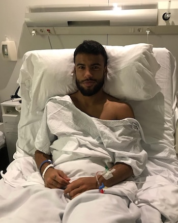 Homem barbudo deitado em uma cama de hospital, vestindo uma camisola e bandagens no braço direito, olhando para a câmera