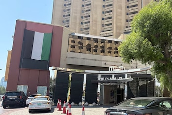 Fachada de un edificio con un cartel que dice "THE PLAZA INN", una bandera vertical de Emiratos Árabes Unidos, varios vehículos, conos y un árbol