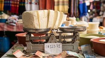 Gran bloque de queso blanco nicaragüense en rodajas sobre una balanza metálica antigua. Billetes de córdobas y monedas dispersos con una etiqueta C$ 123.71. Fondo de mercado.