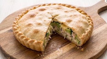 Tarta salada redonda con masa dorada, relleno de pollo deshebrado y brócoli, sobre una tabla de cortar de madera clara. Falta un trozo de la tarta.