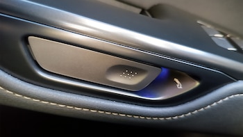 Primer plano de un panel interior de automóvil con un botón metálico, pequeños puntos en relieve, una luz azul y una flecha blanca, sobre cuero negro cosido