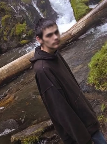 Un joven con sudadera oscura permanece de pie junto a un tronco caído y una cascada en un área boscosa