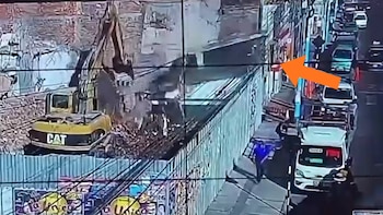 Vista aérea de un sitio de demolición con una excavadora CAT amarilla y escombros; un muro colapsa cerca de una calle con vehículos y personas