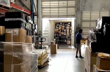 Interior de un almacén mostrando estanterías llenas de cajas y productos embalados; dos hombres trabajan organizando carga, incluyendo sillas de oficina y mesas