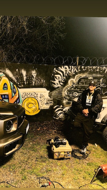 L-Gante posa apoyado en un vehículo blanco, con un maletín de cámara en el suelo y una pared con grafiti de fondo
