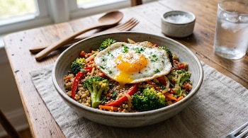 Un tazón cerámico con salteado de quinoa, brócoli, pimiento y un huevo frito sobre una mesa de madera junto a un vaso de agua.