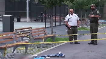 Dos agentes de seguridad uniformados en un parque público, con cinta amarilla de precaución, bancos de madera, cancha de baloncesto y vegetación