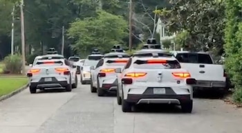 Vista trasera de varios SUVs blancos autónomos en convoy con sensores en el techo, circulando por una calle residencial arbolada