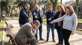 Un hombre sentado en un banco de parque se cubre la cara con las manos. Cinco adultos de pie lo rodean, riendo y apuntándolo. El fondo muestra árboles y caminos.