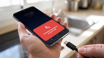 Mano sostiene un celular con pantalla que muestra alerta de "Humedad detectada en el puerto USB" y gotas de agua. Otra mano sostiene un cable cargador con gotas de agua.