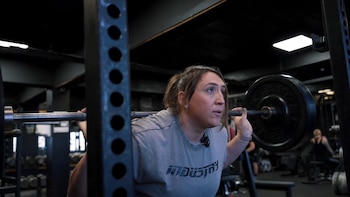 Lucy Underdown sostiene una barra cargada con discos de pesa sobre los hombros dentro de un gimnasio