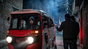 Primer plano de un mototaxi rojo con luces encendidas; el conductor con gorra mira al frente. Al fondo, una persona entrega un pequeño paquete a un ocupante.
