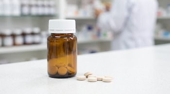Un frasco de vidrio café con tapa blanca y varias tabletas de color claro sobre un mostrador blanco con una farmacia de fondo.