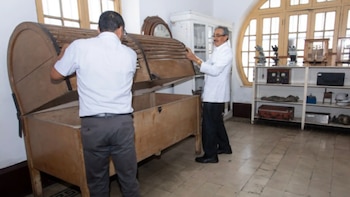 Dos hombres, uno en camisa y otro en bata, abren un baúl de madera con tapa de listones. Detrás hay un estante con objetos y una ventana con rejas