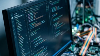 Primerísimo primer plano de una pantalla de computadora inclinada mostrando código ilegible en un editor oscuro. Fondo borroso con componentes electrónicos y un cable plano multicolor