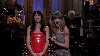 Dakota Johnson con vestido rojo y Taylor Swift con vestido a cuadros permanecen sobre un escenario con músicos al fondo