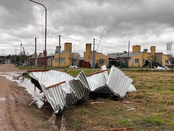 Techo de chapa retorcido en el suelo con charcos de agua, pasto y tierra. Al fondo, casas de un piso, postes de luz y cielo nublado