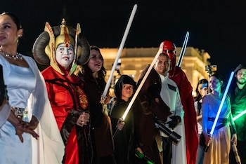Varias personas disfrazadas de personajes de Star Wars como Padmé Amidala, Jedi y un Guardia Imperial, sostienen sables de luz brillando en la oscuridad