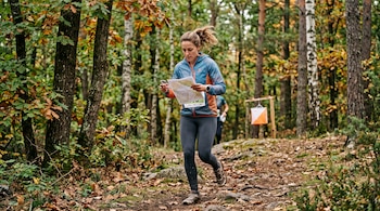 Una mujer sostiene un mapa en un bosque con hojas secas mientras corre hacia un marcador blanco y naranja.
