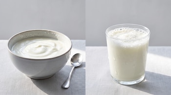 Un bowl de yogur blanco con una cuchara plateada a la izquierda, y un vaso de kéfir blanco con espuma a la derecha sobre una tela clara.