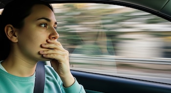Primer plano de una mujer de perfil, con camiseta verde azulado y cinturón de seguridad, mirando por la ventana de un coche en movimiento con la mano cubriendo su boca.