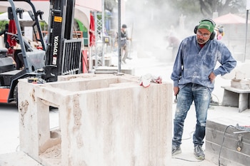 Hombre con gafas, orejeras y bandana, cubierto de polvo, de pie junto a un bloque de piedra en proceso de talla. Guantes y herramientas sobre el bloque. Grúa Toyota en el fondo