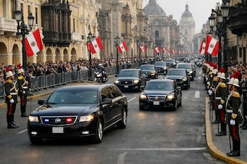 Una caravana de autos negros avanza por una calle flanqueada por militares de gala, público detrás de vallas, edificios coloniales y banderas de Perú.