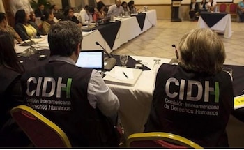 Dos personas de espaldas con chalecos institucionales sentadas frente a una hilera de mesas con computadores y varios asistentes en una sala