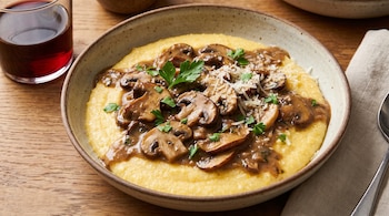 Plato de polenta amarilla con salsa de champiñones marrones, perejil verde y queso blanco rallado. Hay un vaso con vino tinto y una servilleta.