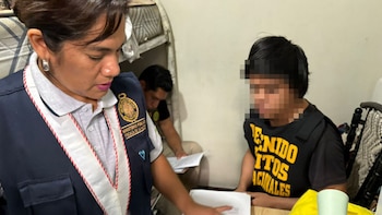 Primer plano de una mujer de uniforme del Ministerio Público mirando documentos, con un hombre de camiseta negra borroso al frente y otro hombre detrás