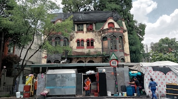 Ilustración de fachada de edificio con ventanas arqueadas, balcón y techo cubierto. Debajo hay puestos de metal, personas, árboles y señales de tráfico.