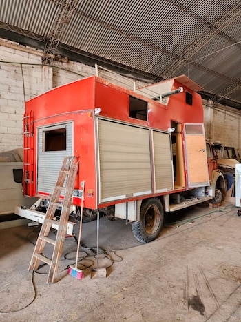 Camión de bomberos rojo en un taller, con aberturas en la carrocería y una escalera de madera apoyada en la parte trasera. Piso de concreto