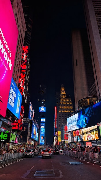Calle urbana de noche con edificios altos, grandes pantallas publicitarias brillantes, vehículos en la calzada y numerosas personas en las aceras