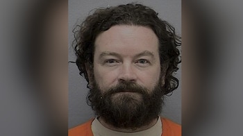 Retrato frontal de un hombre con cabello rizado oscuro y barba espesa, que viste un uniforme presidiario naranja frente a un fondo liso